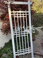 Photo of a wooden trellis painted light grey, showing rectangular lattice of vertical and horizontal slats, standing outdoors near a tree and fence.