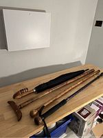 Photo showing one black umbrella with carved wooden handle, three carved wooden canes, and one black collapsible cane with foam grip arranged on a table.