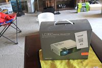 Photo showing Apeman LC350 digital projector boxed with handle on top, placed on a yellow cloth over a table, in indoor setting with industrial background.