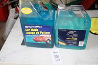 Two gallon-sized car wash bottles on a table, one Turtle Wax Car Wash and one Ice Finish Care Wash Nettoyant, both containing blue-colored liquids.