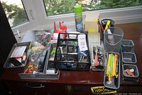 Overview of the assortment of office items spread on wooden desk including organizers, pens, pins, and cleaning spray.