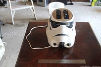 Side angle view of Stormtrooper helmet toaster on wooden table showing power cord and toaster slots.