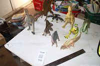 Photo showing multiple plastic dinosaur toy figures on a white table, arranged with some larger behind smaller toys.