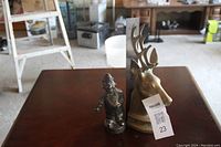 Two brass sculptures on a wooden table: a deer head bust and a smaller man figurine with a ruler for height reference.