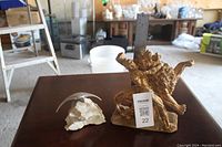 Photo shows two sculptures on a wooden surface with a ruler for scale; golden animal sculpture and silver bird sculpture on rock base.