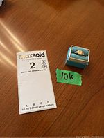 Top down photo showing the small blue jewelry box containing the gold ring with monogram next to a MaxSold auction note card and green tape labeled '10K'.