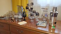 Assorted silver overlay glassware including stemmed glasses, pitchers with floral silver overlay, and a silver condiment set arranged on a wooden dresser with a yellow curtain background.