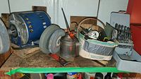 Bench grinder with blue motor housing and one grinding wheel attached plus extra wheel on the table near a metal oil can and a box with assorted small tools.