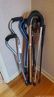 Photo shows six mobility canes leaning against a wall. Three are aluminum with ergonomic black foam handles, including one blue colored cane. Three are wooden with traditional curved handles and rubber cane tips.