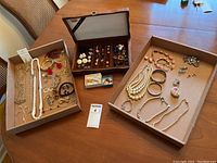 Three boxes and a wooden case containing assorted costume jewelry including multiple necklaces, bracelets, earrings, brooches, and rings arranged on a table.