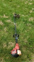Full view of Homelite gas trimmer lying on grass showing red engine housing, black shaft and handle, and cutting guard at head.