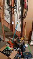 Photo showing a basement area with tools and hardware stored by stairs, including a carry bucket with gloves and various hand tools like hammers and screwdrivers, and cardboard boxes with hardware items on the floor.