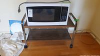 White Hamilton Beach microwave on a metal and wood two-tier microwave cart with wheels.