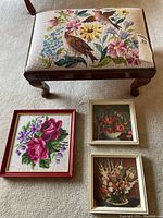 Overview of footstool with needlepoint top and three framed needlepoint pieces laid on floor