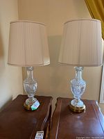 Pair of crystal table lamps displayed on wooden side tables, showing the full lamps including crystal bases, brass bottom bases, and white pleated shades.