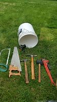 Photo of all tools laid out on grass showing a hand saw, hammer, mallet, pipe wrench, and a white bucket containing a clip-on vise.