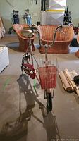Front view of vintage folding bike showing basket, handlebars, and seat with concrete basement floor and wicker furniture background.