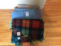 Three wool blankets and a quilt stacked on the floor. The Buchanan tartan blanket shows a multicolor check pattern with fringe. The Kenwood blanket is light blue with a satin edge. A patterned quilt is rolled in the background.