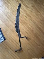 Top view of the antique hand forged iron saw laid flat on a hardwood floor showing the entire curved blade with large teeth and crank handle.