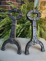 Front view of pair of solid brass Arts and Crafts style firedogs with visible rivet details and original patina, placed outdoors against green foliage background.