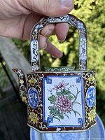 View of hexagonal hand-painted enamel teapot held, showing floral motifs and geometric patterns in multiple colors with brass handle.