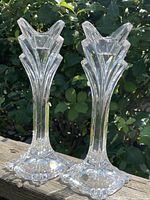 Pair of 9.5 inch tall clear crystal candleholders with flared stylized tops standing outdoors with a green leafy background.