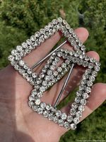 Pair of rectangular shoe buckles held in hand, showing full view with clear sparkling stones and metal bars at the back.
