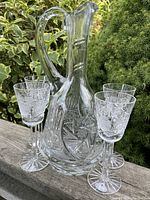Photo showing the crystal decanter with handle and four matching crystal stems, placed outdoors on a wooden surface with greenery in the background.