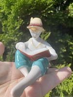 Front view of the porcelain figurine showing the boy in a hat reading a book, seated with bent knees, painted in beige, white, turquoise, and reddish-brown.