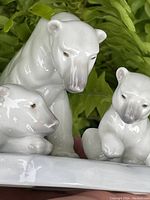 Close-up of three white porcelain polar bears, showing detailed facial and body features with glossy finish against green foliage background.