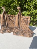 Front angle of pair of bookends shaped as sailing ships in metal with a bronze finish placed outdoors on a white surface with green bushes in background.