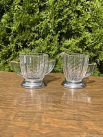Two-piece Art Deco cream and sugar set outdoors on table showing pressed glass texture and silver-tone metal bases.