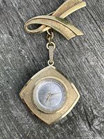 Front view of gold-tone watch pin attached to decorative ribbon-shaped pin, showing square watch case with clear faceted crystal and silver dial with gold hands and indices.
