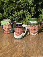 Three English Toby mugs displayed outdoors on wood, with green foliage background; mugs include Sairy Gamp (green bonnet), Tony Eweller (black top hat), and Raleigh (green hat with feather).