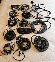 Photo showing a collection of different lengths and types of black audio cables coiled on a light surface. Cables include 1/4 inch plugs, RCA connectors, and miscellaneous instrument cables.