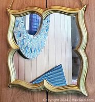 Close-up view showing the square mirror with wavy scalloped gold frame reflecting part of the room.