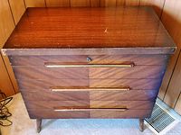 Front view of the cedar chest showing wood grain finish and three horizontal gold-plated insets on the drawers, solid tapered legs visible.