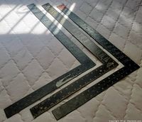 Five solid metal L-shape carpenter squares with black finish and ruler measurements laid out on a quilted surface, showing signs of use.