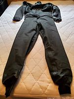 Full length front view of black Ultra drysuit with white shoulder stripes lying flat on quilted surface showing legs, torso, and arms.