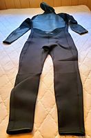 Photo showing the full length of the black wetsuit laid flat with hood visible, highlighting the form-fitting shape covering torso, arms, legs, and head.