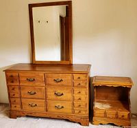 Front view of solid wood dresser with attached rectangular mirror, multiple drawers with metal handles. Bedside table beside it showing wear on open shelf and drawer fronts.