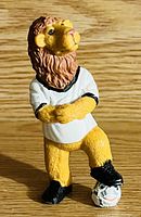 Front view of Goleo mascot figurine, showing yellow lion character with brown mane, white jersey, black shoes and soccer ball under foot.