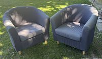 Two gray upholstered IKEA club chairs placed on a grass lawn, showing front and side views. Each chair has a curved back and armrests, wooden legs, and a removable cushion seat.