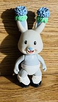 Photo of a plastic bunny figurine with blue flowers decorating the tops of its ears, expressive face, light blue torso, white shoes and diaper-like bottom.