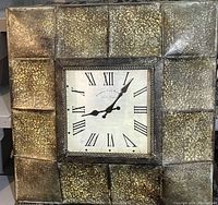 Front view of the large metal wall clock showing textured metallic frame and square face with Roman numerals.