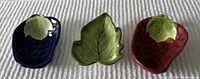 Three ceramic bowls shaped as a blue blackberry, green leaf, and red strawberry with leaf accents, placed side by side on a white textured surface.