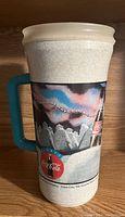 Side view of vintage Coca-Cola thermos cup showing colorful mountain and Coca-Cola bottle artwork and blue handle.