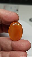 Close-up photo of a natural mandarin orange carnelian gemstone held between fingers, showing an oval cabochon cut with smooth glossy surface