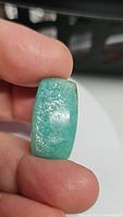 Close-up front view of cushion cut teal natural amazonite gemstone held between thumb and index finger showing polished glossy surface and natural inclusions.