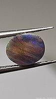 Close-up of the oval cabochon black opal held by metal tweezers showing color flashes and surface condition.